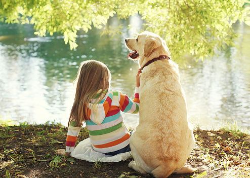 Little girl and yellow labrador retriever sitting by lake or river
