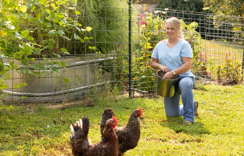 A women feed two chickens in the yard with a garden full of flowers and herbs behind her. This represents how poultry gut health is supported by Nutrena NatureWise feeds with FlockShield and Herbal Blend.